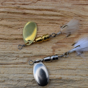 May include: Two fishing lures with white feathers and silver and gold metal bodies. The lures are on a wooden surface.