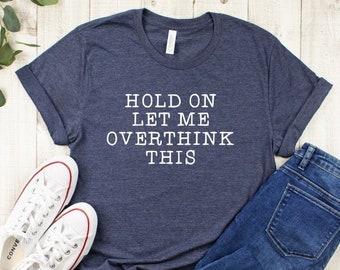 May include: A navy blue t-shirt with the text "HOLD ON LET ME OVERTHINK THIS" in white, paired with blue jeans and white canvas sneakers with red accents. The items are arranged on a light wood surface.