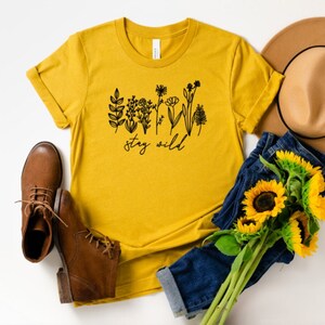 May include: A mustard yellow t-shirt with a black graphic of wildflowers and the text "stay wild". The shirt is styled with a brown hat, blue jeans, and brown boots.