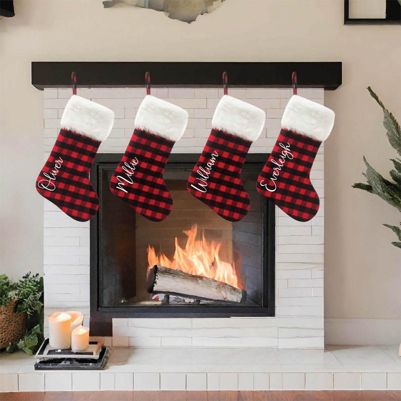 May include: Four red and black buffalo plaid Christmas stockings with white faux fur trim hanging on a mantel above a fireplace. The stockings are personalized with the names Oliver, Millie, William, and Everleigh.