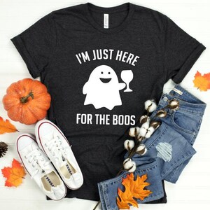 May include: Dark gray t-shirt with the text "I'M JUST HERE FOR THE BOOS" and a white ghost holding a wine glass. The shirt is paired with blue denim shorts, white sneakers, and fall-themed decorations including a pumpkin and leaves.
