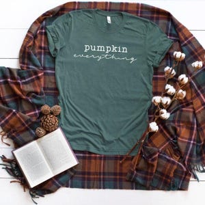 May include: A teal t-shirt with the words "pumpkin everything" in white script is displayed on a white wooden surface. A plaid blanket in shades of brown, green, and purple surrounds the shirt. A book, pinecones, and cotton stems complete the fall-themed arrangement.
