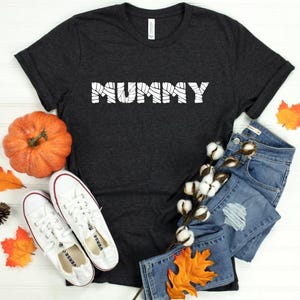May include: A dark gray t-shirt with the word "MUMMY" in white, styled with a pumpkin, white sneakers, denim shorts, and fall foliage. Perfect for Halloween or fall festivities.