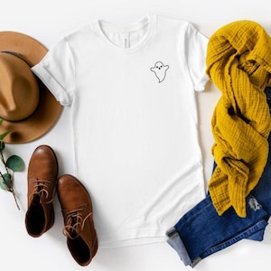 May include: A white t-shirt with a small black ghost graphic, paired with brown boots, a tan hat, blue jeans, and a mustard yellow scarf. The items are arranged on a white surface, creating a casual, fall-themed aesthetic.