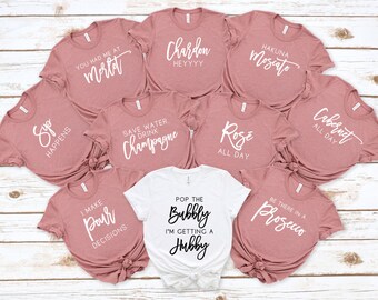 May include: A collection of dusty rose and white t-shirts with wine-themed phrases like "Hakuna Moscato" and "Cabernet All Day." One white shirt reads "Pop the Bubbly I'm Getting a Hubby."