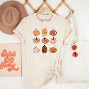 May include: Cream-colored t-shirt featuring nine cartoon pumpkins in various colors and designs. A canvas tote bag with red sunglasses hangs to the right. A framed print with the words "Sweet Life Honey" is on the left.
