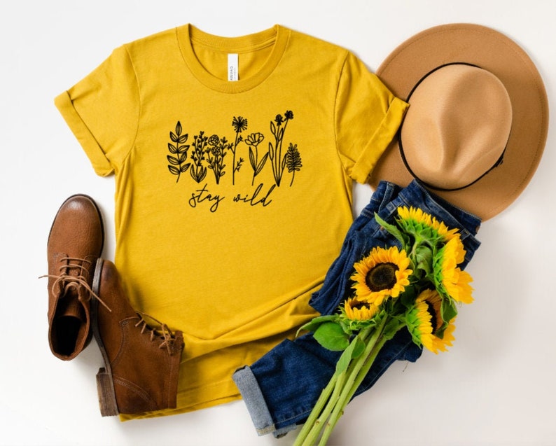May include: A mustard yellow t-shirt with a black graphic of wildflowers and the text "stay wild". The shirt is styled with a brown hat, blue jeans, and brown boots.
