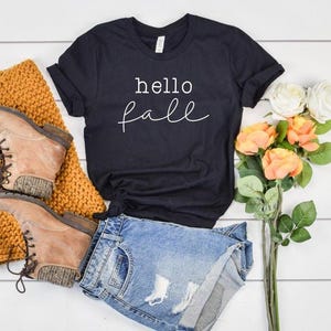 May include: A black t-shirt with the words "hello fall" in white script, paired with denim shorts, brown boots, and a mustard-colored scarf. A bouquet of orange and white flowers adds a touch of color.