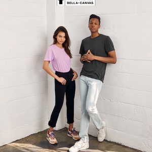 May include: Two people wearing t-shirts and jeans stand in a corner of a room. The woman is wearing a light pink t-shirt and black jeans. The man is wearing a dark gray t-shirt and light blue jeans. The text "WE PROUDLY USE BELLA+CANVAS" is in a white box in the upper right corner of the image.