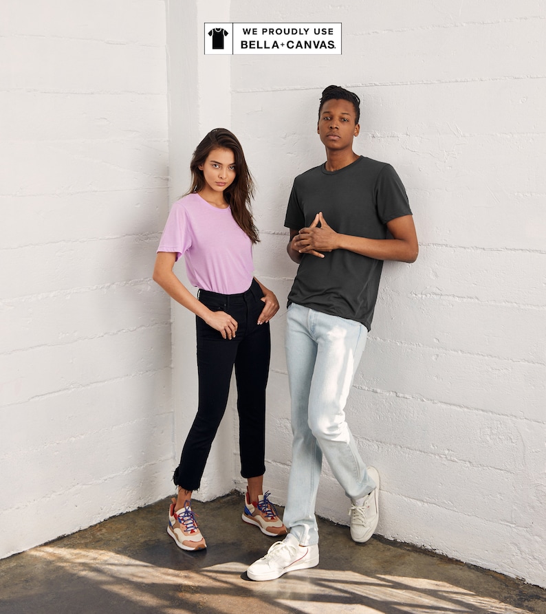 May include: Two people wearing t-shirts and jeans stand in a corner of a room. The woman is wearing a light pink t-shirt and black jeans. The man is wearing a dark gray t-shirt and light blue jeans. The text "WE PROUDLY USE BELLA+CANVAS" is in a white box in the upper right corner of the image.