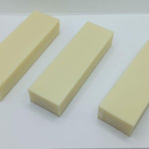 May include: Three rectangular bars of pale yellow soap are arranged on a white surface. The bars have a smooth, solid appearance, with a slightly angled top. The soap bars are evenly spaced, creating a clean and simple composition.