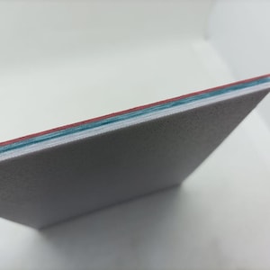 Puede incluir: Primer plano del borde de una hoja blanca texturizada con tres capas de material coloreado visibles: una capa roja fina, una capa azul fina y una capa blanca más gruesa.