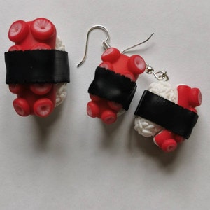 K&ouml;nnte beinhalten: Ein Paar H&auml;ngeohrringe mit einem Sushi-inspirierten Design mit roten Krakenarmen und wei&szlig;em Reis, gekr&ouml;nt mit einem schwarzen Seetangstreifen. Die Ohrringe sind aus Polymer Clay gefertigt und haben silberne Haken.