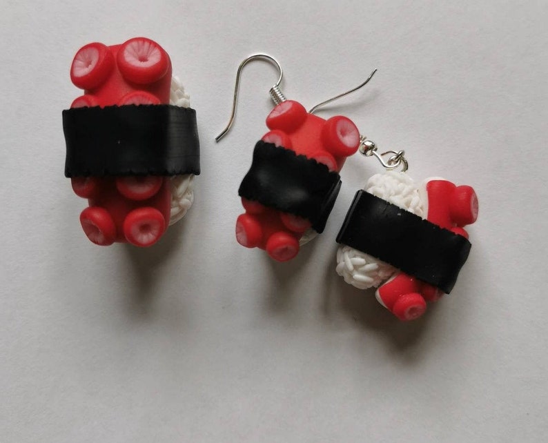 K&ouml;nnte beinhalten: Ein Paar H&auml;ngeohrringe mit einem Sushi-inspirierten Design mit roten Krakenarmen und wei&szlig;em Reis, gekr&ouml;nt mit einem schwarzen Seetangstreifen. Die Ohrringe sind aus Polymer Clay gefertigt und haben silberne Haken.