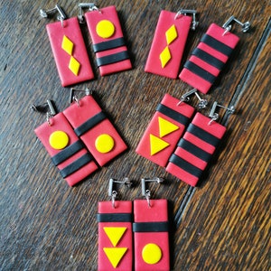 May include: Six pairs of red and black polymer clay earrings with yellow geometric shapes. The earrings are in the shape of a rectangle with a black stripe across the middle. The geometric shapes include a diamond, a circle, and a triangle.