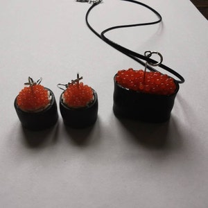 K&ouml;nnte beinhalten: Ein Set an Sushi-Schmuck, bestehend aus einer Halskette und Ohrringen. Die Halskette hat einen schwarzen und roten Sushi-Rollen-Anh&auml;nger mit rotem Kaviar obenauf. Die Ohrringe sind passende Sushi-Rollen mit rotem Kaviar.