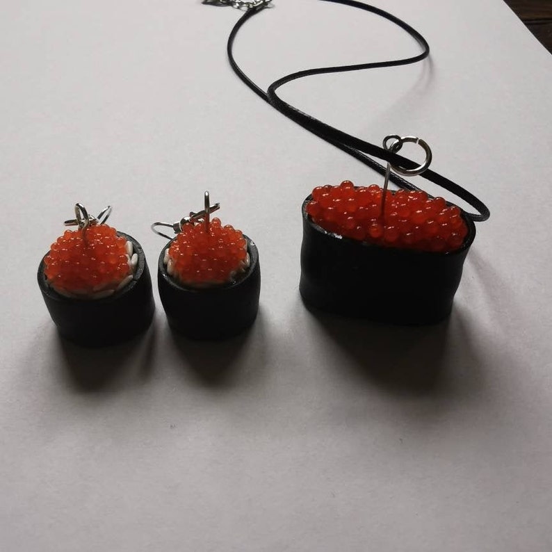 K&ouml;nnte beinhalten: Ein Set an Sushi-Schmuck, bestehend aus einer Halskette und Ohrringen. Die Halskette hat einen schwarzen und roten Sushi-Rollen-Anh&auml;nger mit rotem Kaviar obenauf. Die Ohrringe sind passende Sushi-Rollen mit rotem Kaviar.