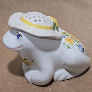 Puede incluir: Figura de rana de cerámica blanca con sombrero blanco con banda amarilla y detalles florales. La rana tiene ojos azules y está decorada con motivos florales amarillos y azules. Un artículo decorativo.