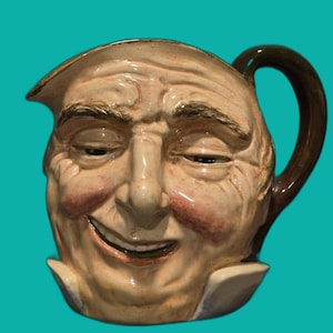 May include: A ceramic jug in the shape of a smiling man's face. The jug features a brown handle and spout, with a cream-coloured face, rosy cheeks, and dark brown eyebrows. The man's face has a cheerful expression.