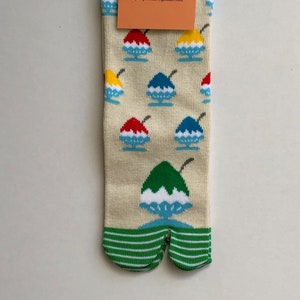 May include: A pair of cream-colored tabi socks with a split toe. The socks feature a repeating pattern of colorful shaved ice with a cherry on top. The socks have a green stripe at the top of the toe split. The socks have a tag that reads "flop-socks (because it's 50' somewhere) myflopsocks@gmail.com"