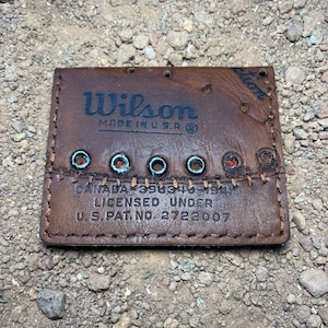 Puede incluir: Una etiqueta de cuero marrón con el logotipo de Wilson y "MADE IN U.S.A." impreso en negro. La etiqueta tiene ojales metálicos y el texto "CANADA 398346 1941 LICENSED UNDER U.S. PAT NO. 2722007".