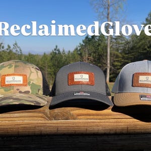 Op de afbeelding: Drie baseballpetten met de tekst "ReclaimedGlove" erboven. De petten hebben een leren patch met de merknaam. Eén pet is camouflage, één is grijs en één is lichtgrijs met een bruin vizier.