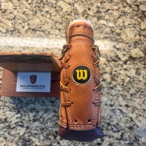 Op de afbeelding: Een bruine leren flessenhouder gemaakt van een honkbalhandschoen, met het Wilson-logo in geel en zwart. De houder heeft witte stiksels en een donkerbruine basis. De tekst "RECLAIMEDGLOVE" is zichtbaar op een klein bord.