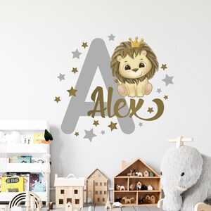 May include: A gray and gold wall decal featuring a lion wearing a crown, the letter A, and the name Alex. The decal is surrounded by gold stars.