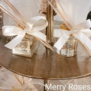 May include: Two glass bottles of aromatherapy diffuser oil with white ribbon bows and wooden sticks. The bottles are labelled "Luxury Zen Aromatherapy" and have gold lids. The bottles are on a gold metal stand.