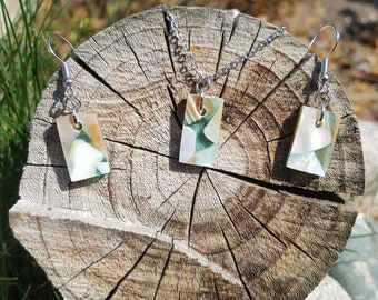Puede incluir: Un conjunto de joyería compuesto por un collar y dos pendientes a juego, cada uno con un colgante rectangular hecho de fragmentos de concha iridiscente en tonos blanco, verde y bronceado, suspendido de ganchos de metal plateado y una cadena.