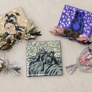 May include: Three square tiles featuring black dog illustrations and metallic foil accents, paired with small dried floral arrangements. One tile shows a person with a dog, while two others depict dogs resting. Two additional dried flower bouquets are wrapped in clear plastic.