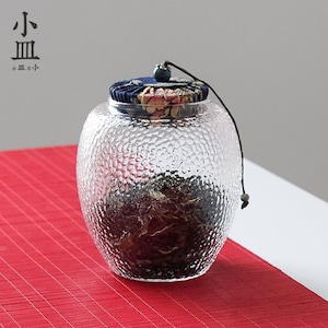 May include: A clear glass jar with a textured surface and a fabric lid with floral and blue patterns. The jar is filled with loose tea leaves and has a small green bead on the lid.
