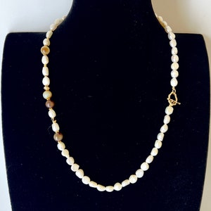 Könnte beinhalten: Eine Perlenkette mit goldenem Verschluss und Akzenten. Die Halskette hat weiße, oval geformte Perlen und einige braune Perlen. Die Halskette wird auf einer schwarzen Schaufensterpuppe präsentiert.