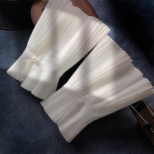 May include: A pair of white, pleated, flared sleeve cuffs featuring a horizontal fabric band across the centre. One cuff is accented with a single, small, round white pearl button attached to the band, while the other remains plain.