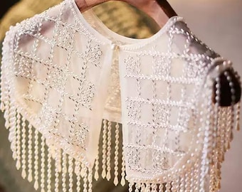 May include: Ivory-colored, sheer lace collar with intricate floral embroidery and dangling pearl bead fringe. The collar is displayed on a wooden hanger, showcasing its delicate design and texture. A stylish accessory for adding elegance to any outfit.