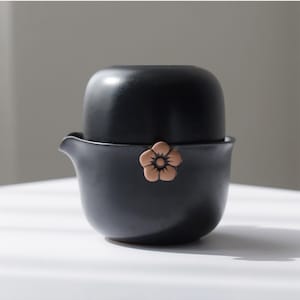 以下が含まれることがあります： 黒いセラミックティーセット。蓋と注ぎ口付き。ティーポットの側面には、小さなベージュ色の花の装飾が施されています。お茶を淹れて提供するために設計されたセットです。