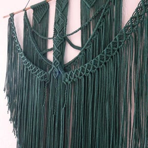 Op de afbeelding: Een handgemaakte macramé wandhanger in diep bosgroen. Het textielkunstwerk heeft ingewikkelde knoopmotieven en lange, vloeiende franjes, opgehangen aan een natuurlijke houten deuvel. Dit decoratieve item voegt een bohemien touch toe aan elke interieurruimte.