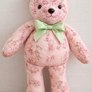 May include: A plush teddy bear with a pink floral pattern. The bear has a black nose, black eyes, and a light green bow. The bear is standing upright and is centered in the frame.