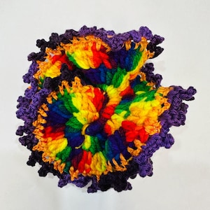 May include: A handmade, colorful crochet doily. The center features a vibrant rainbow of colors, including red, orange, yellow, green, blue, and purple. The edges are trimmed with a dark purple and black scalloped border, adding a decorative touch.