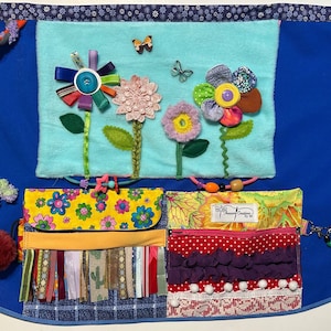 May include: A blue sensory apron with various textures and embellishments. Features include fabric flowers, ribbons, buttons, and small pockets. The apron has a blue background with a floral design and a pom-pom.