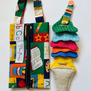 May include: A colorful fabric zipper pouch with a school-themed print, including a bus, pencils, and a book. The pouch has a zipper closure and a fabric handle. A separate fabric toy shaped like an ice cream cone with colorful layers and a button on top.