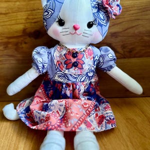 May include: A plush cat doll with white fabric skin, black button eyes, and long eyelashes. The cat wears a blue and white floral patterned headpiece with a matching fabric flower, a gold-tone necklace, and a dress featuring blue and coral floral prints.