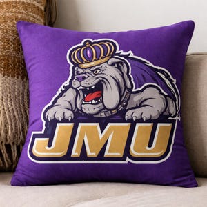 May include: Purple decorative pillow featuring a bulldog wearing a crown, with the letters "JMU" in gold. The bulldog is gray and white with an open mouth and red tongue. The pillow is on a beige couch.