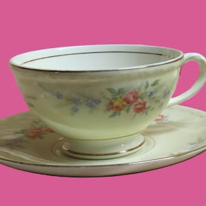May include: Two vintage teacups and saucers. The cups and saucers are cream-colored with a floral design in pink, yellow, and blue. Each piece has a gold trim around the edges. The teacups have a handle and sit on a pedestal base.