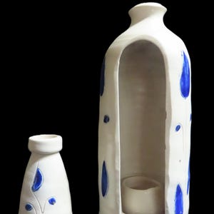 May include: Two white ceramic vases with blue accents. The larger vase has a tall, open design with a small, round interior shelf. The smaller vase is bottle-shaped. Both vases feature blue teardrop and dot patterns.