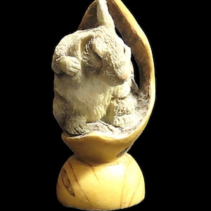 May include: A cream-colored rabbit figurine sculpted inside a cracked eggshell. The rabbit is detailed with fur texture and a pink nose. The eggshell is a light yellow, and the base is a rounded shape. The background is black.