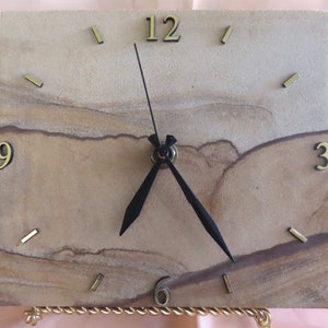 May include: A square sandstone clock with black hands and gold Roman numeral hour markers. The clock is on a gold stand.