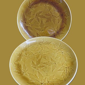 May include: Two ceramic plates with a leaf design. The plates are a warm yellow-brown color with a glossy finish. The leaf pattern is embossed, creating a textured surface. The plates have a white rim.