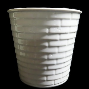 May include: A white ceramic vase with a textured brick pattern. The cylindrical vase has a slightly flared top and is set against a black background. The vase is likely used for holding flowers or as a decorative item.