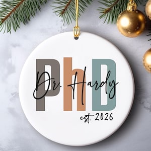 May include: White ceramic Christmas ornament with the text "PhD Dr. Hardy est. 2026" in a modern font. The letters "PhD" are in gray, orange, and teal. The ornament hangs from a gold string, with gold ornaments and evergreen branches in the background.